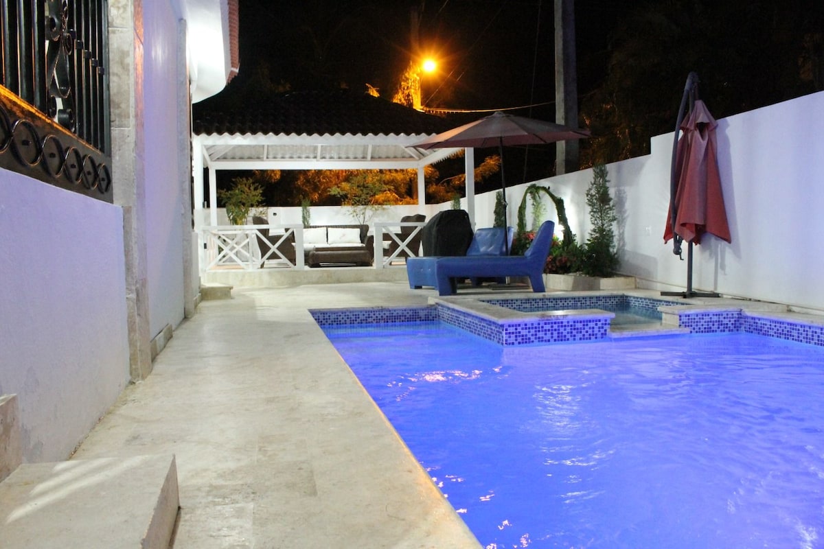 A modern pool area is illuminated by soft blue lighting, complemented by a relaxing lounge space. Nearby, a covered gazebo offers seating and features a light-colored sofa. The surrounding walls are painted white, enhancing the sense of openness in the outdoor space.