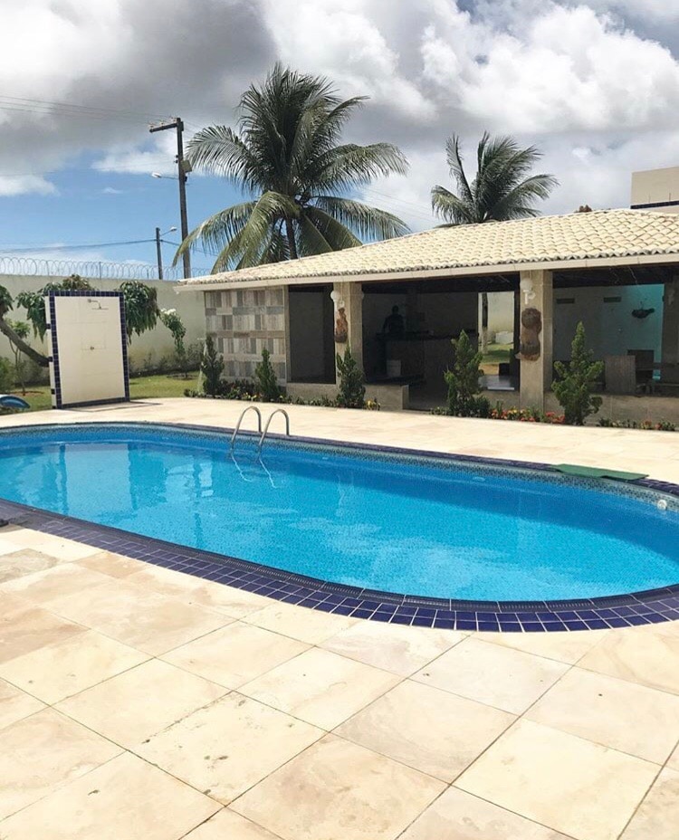 A clear swimming pool is framed by tiled decking, surrounded by lush greenery and palm trees. An outdoor well-shaded area features a structure with seating, providing a relaxed environment for gatherings. The sky is partly cloudy, creating a bright atmosphere.