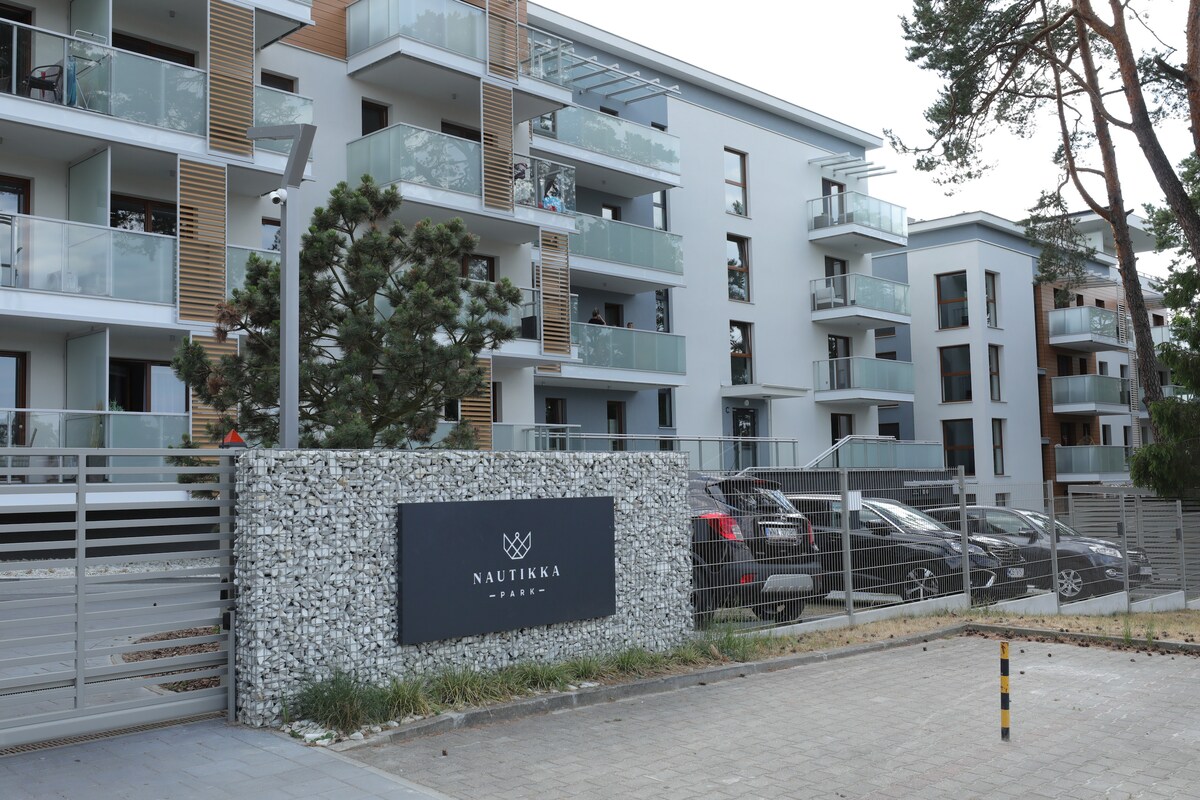 The building is characterized by modern architecture with multiple balconies featuring glass railings. A stone-clad sign displaying 'NAUTIKKA' is prominently placed at the entrance. The surrounding area includes landscaped greenery and a gated entrance providing access to a parking area.