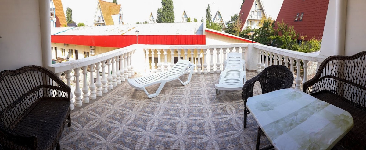 A spacious balcony is featured, equipped with two lounge chairs and a wicker seating area. A small table rests on the patterned tile floor, enhancing the outdoor space surrounded by a white balustrade. Nearby structures and greenery are visible in the background.