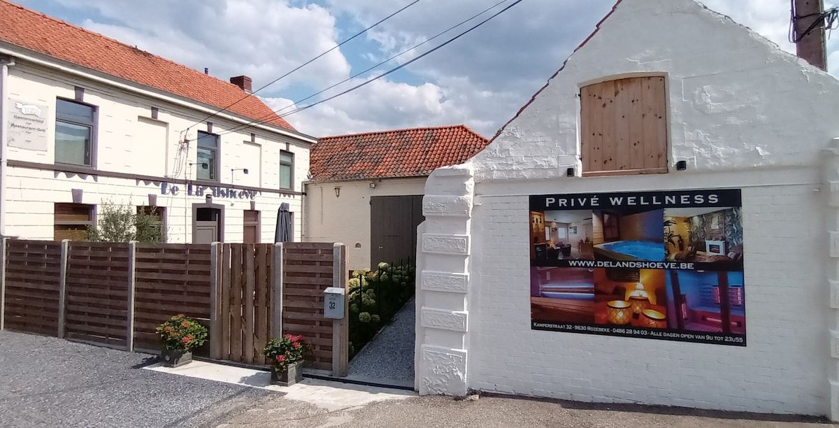A charming exterior view of a white-walled building with a wooden gate, bordered by flowers. The sign for the private wellness area is prominently displayed, indicating its availability for guests. The structure's traditional architecture contrasts with the vibrant sky overhead.