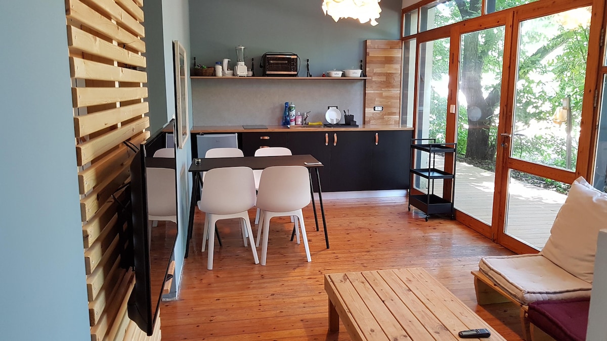 An open living area features wooden flooring and a cozy dining space with a table and four chairs. A kitchenette is equipped with essential appliances, including a microwave and a refrigerator. Large sliding doors provide natural light and reveal the surrounding greenery.