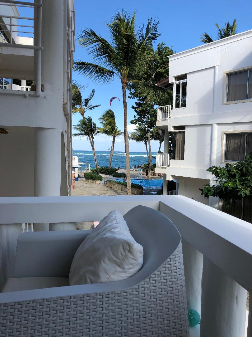 A serene balcony view showcases a white chair with a soft pillow. Palms sway gently in the breeze, leading down to a calm ocean. A glimpse of a pool and the beach can be seen, hinting at a peaceful outdoor atmosphere.