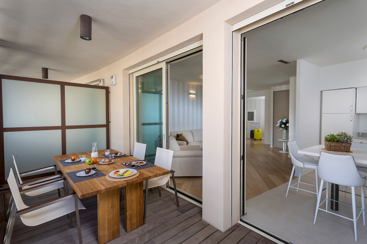 An outdoor terrace is furnished with a wooden dining table surrounded by white chairs. The glass doors lead to a well-lit living area, enhancing the connection between indoor and outdoor spaces. A cozy atmosphere is created by the simplicity of the furnishings.