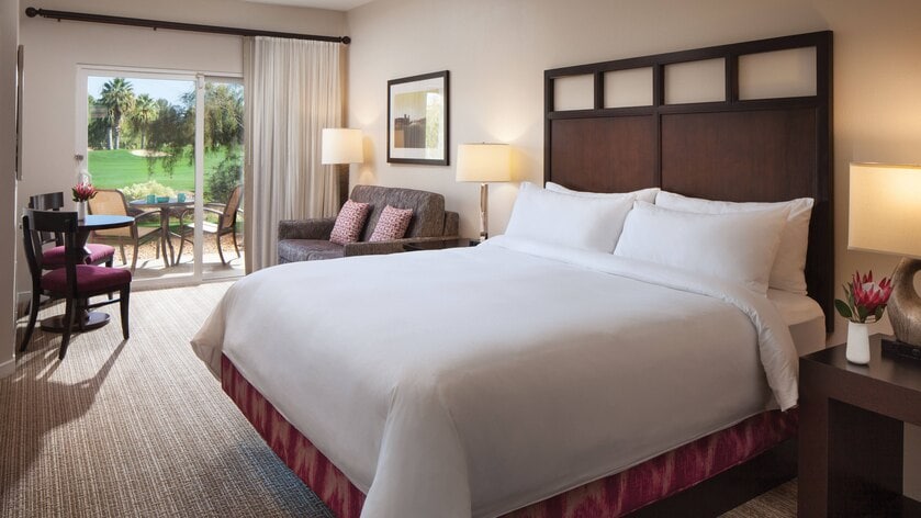 A spacious studio features a large king bed with crisp white linens. A sofa bed and wooden dining table with chairs are also visible. Bright light streams in from the sliding glass doors, which lead to a furnished patio overlooking lush green scenery.