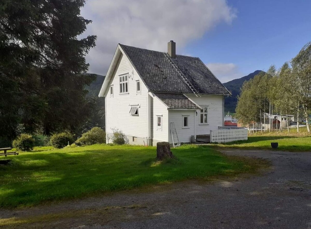 A traditional two-story house is set on a grassy plot, surrounded by trees. The white exterior features multiple windows, and the roof is finished in dark shingles. Natural scenery of rolling hills and trees is visible in the background, offering a serene rural atmosphere.