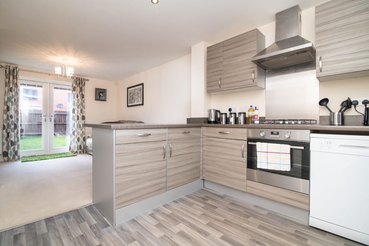 A modern kitchen area is presented with light wood cabinetry and stainless steel appliances. An open-plan layout invites natural light from the glass doors, leading to a grassy outdoor space. Counter space is equipped with various kitchen tools and condiments.