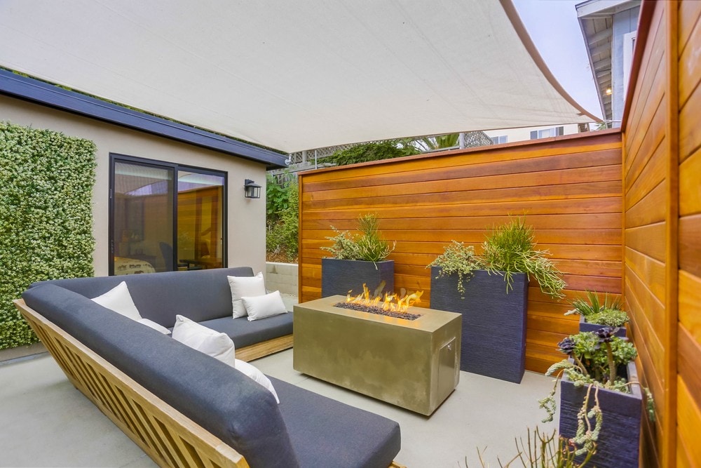An inviting outdoor seating area is furnished with a modern gray sofa and accented by soft white pillows. A sleek fire pit is centered among tall planters filled with greenery, while a fabric canopy provides shade. Natural wood panels create a warm and private environment.