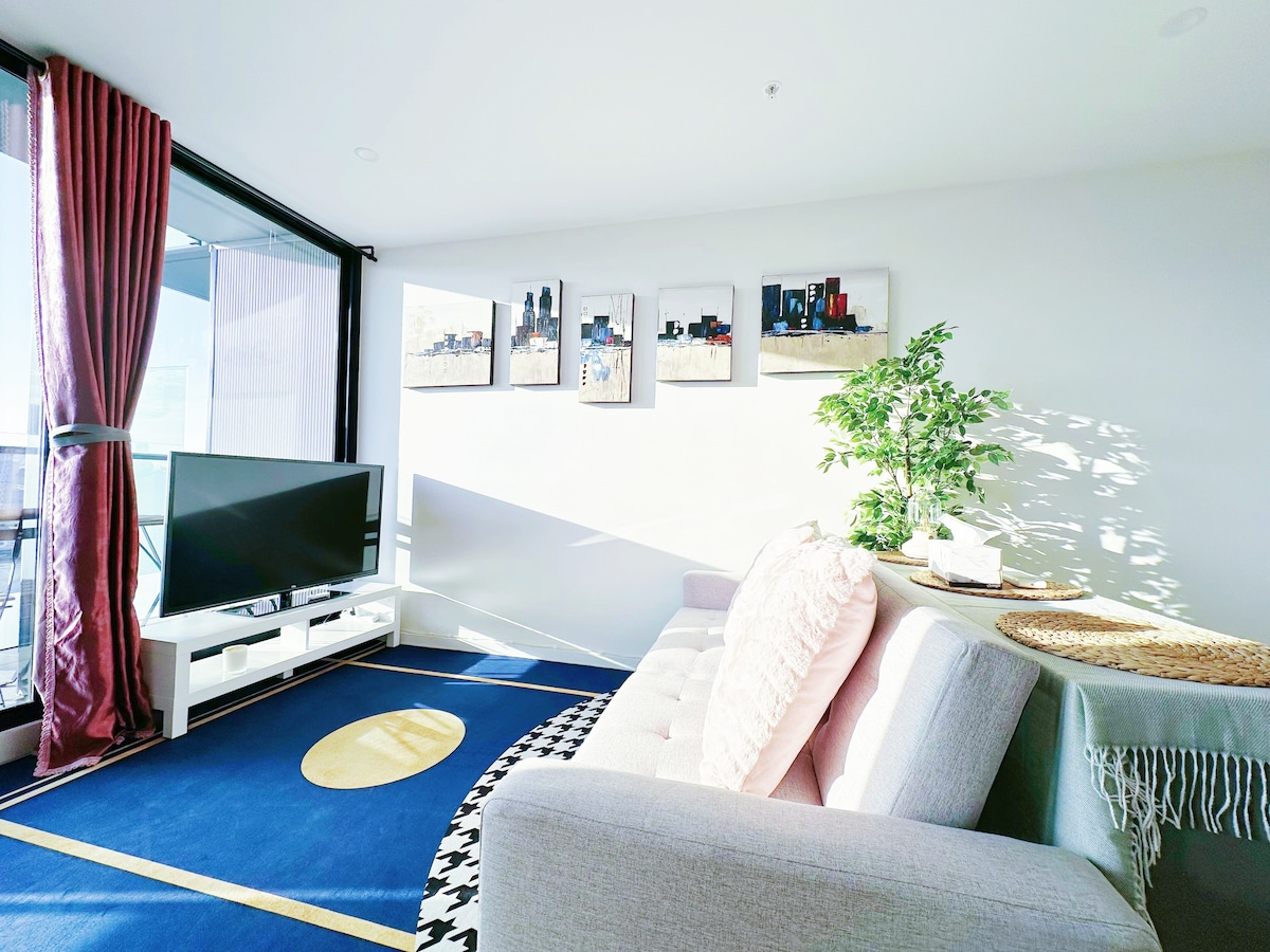 A spacious living area features a large sofa and a flat-screen TV positioned across from a floor-to-ceiling window. Artwork adorns the wall, and natural light casts a bright and airy atmosphere. A decorative plant and a small table add to the welcoming ambiance.