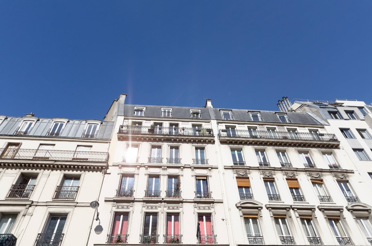 A traditional Parisian building stands under a clear blue sky. The façade features multiple balconies adorned with plants and colorful shutters, contrasting with the light-colored walls. Architectural details highlight the charm of the area, creating a sense of the local atmosphere.