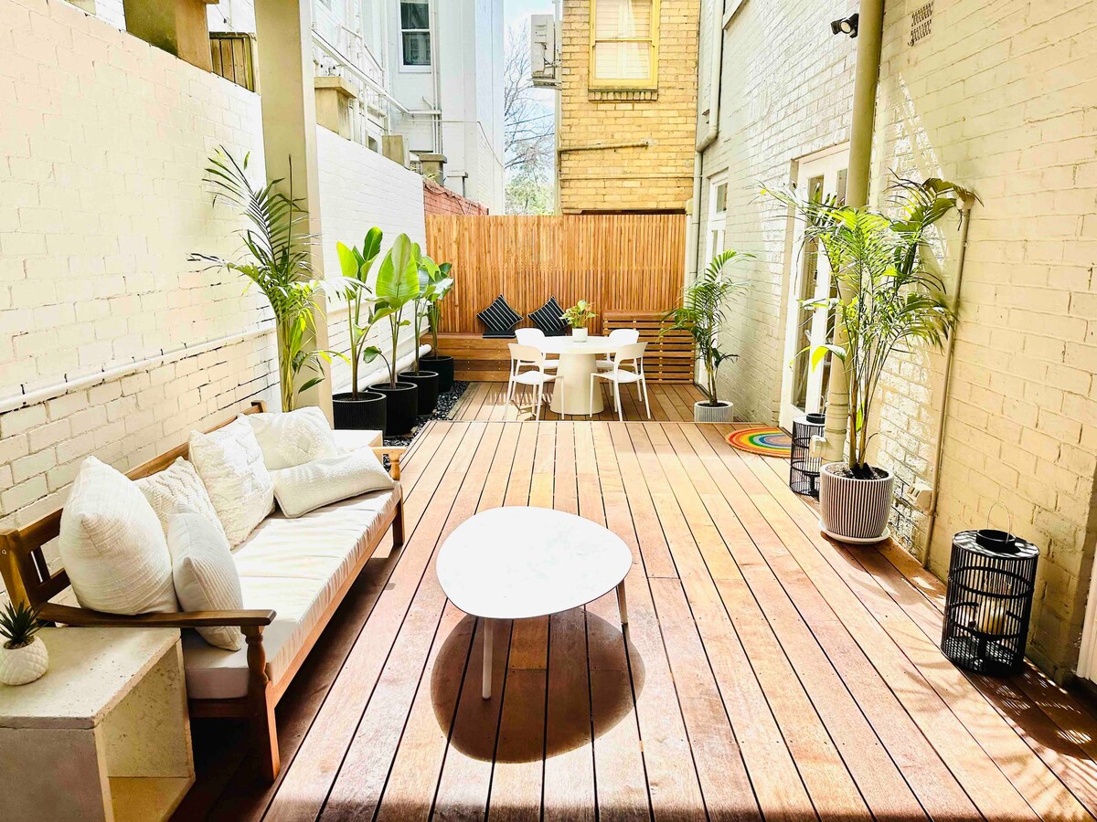 A spacious private courtyard is shown, featuring wooden decking and potted plants. Comfortable seating is arranged around a low table, with a dining area set for meals. Natural light enhances the inviting atmosphere, and decorative elements add a touch of greenery.