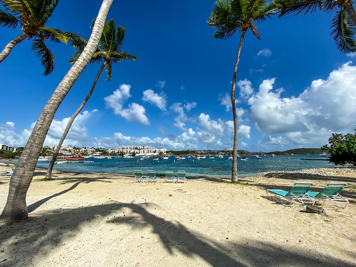 A beach scene features soft sand and gently swaying palm trees. Sun loungers are arranged on the shore, facing the calm water where boats are docked. A backdrop of nearby hills and clouds adds texture to the serene coastal landscape.