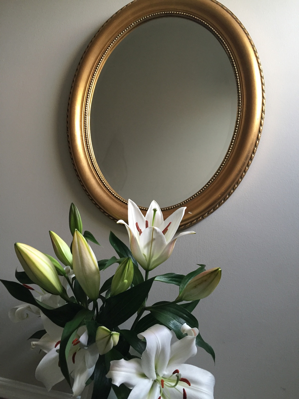 An elegant golden oval mirror is mounted on a soft-colored wall, reflecting ambient light. Below, a bouquet of white lilies, both closed and in full bloom, is arranged with green foliage, adding a touch of nature to the indoor space.