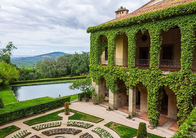An elegant building is surrounded by lush greenery and vines, showcasing archways that frame the view of a serene reflecting pool. The meticulously designed garden features neatly arranged flower beds and stone pathways, enhancing the tranquil outdoor atmosphere.