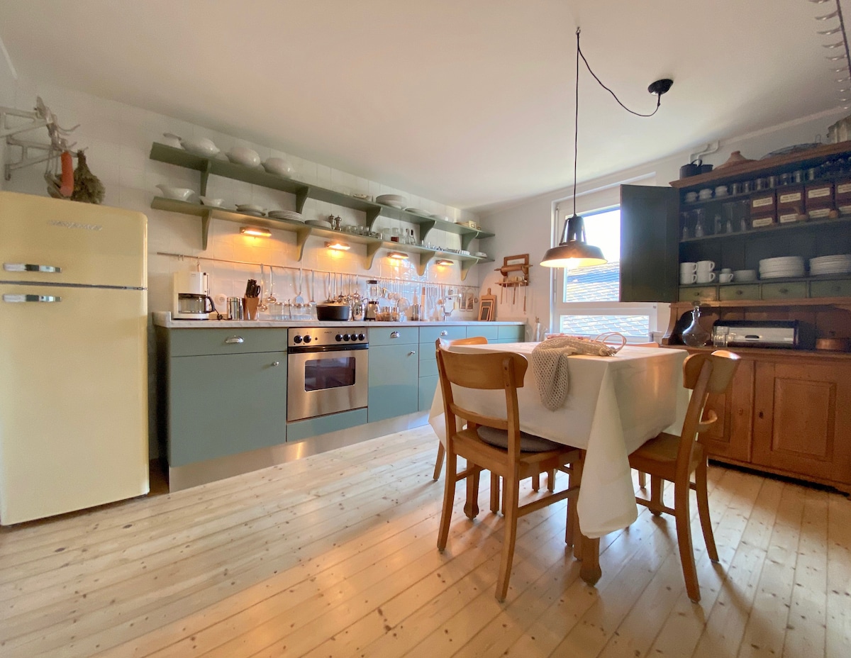 A modern kitchen is presented with seafoam green cabinetry, stainless steel appliances, and a retro-style refrigerator. A wooden dining table is set for four with light-colored linens, while ambient lighting highlights glassware and utensils on open shelves. Natural light enters from a window, enhancing the inviting space.