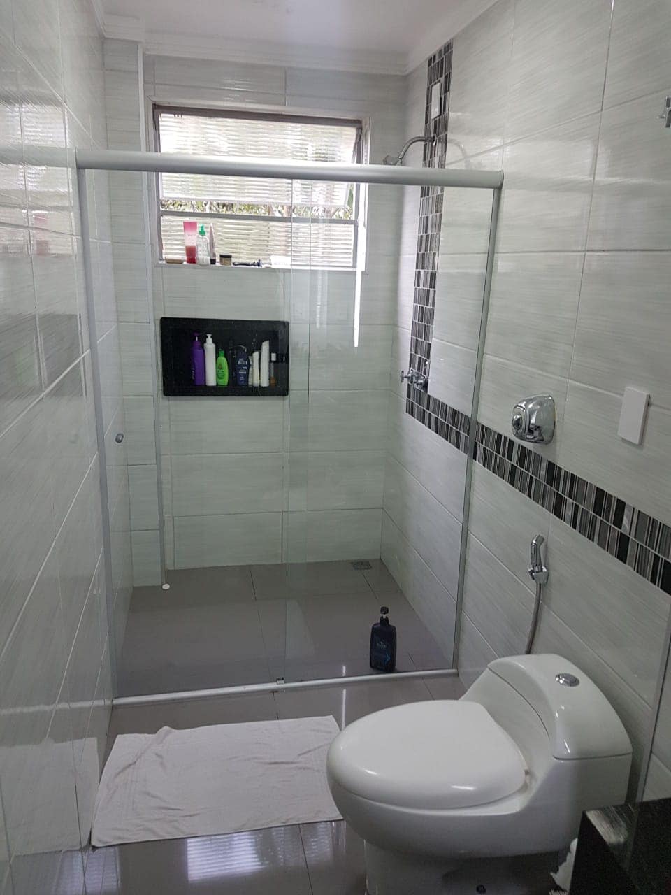 A modern bathroom is depicted, featuring a glass-enclosed shower with a rainfall showerhead. The tiled walls are accented with dark stripes, and amenities are neatly arranged on a shelf. A clean, white toilet is positioned next to the shower area, with a soft mat on the floor.