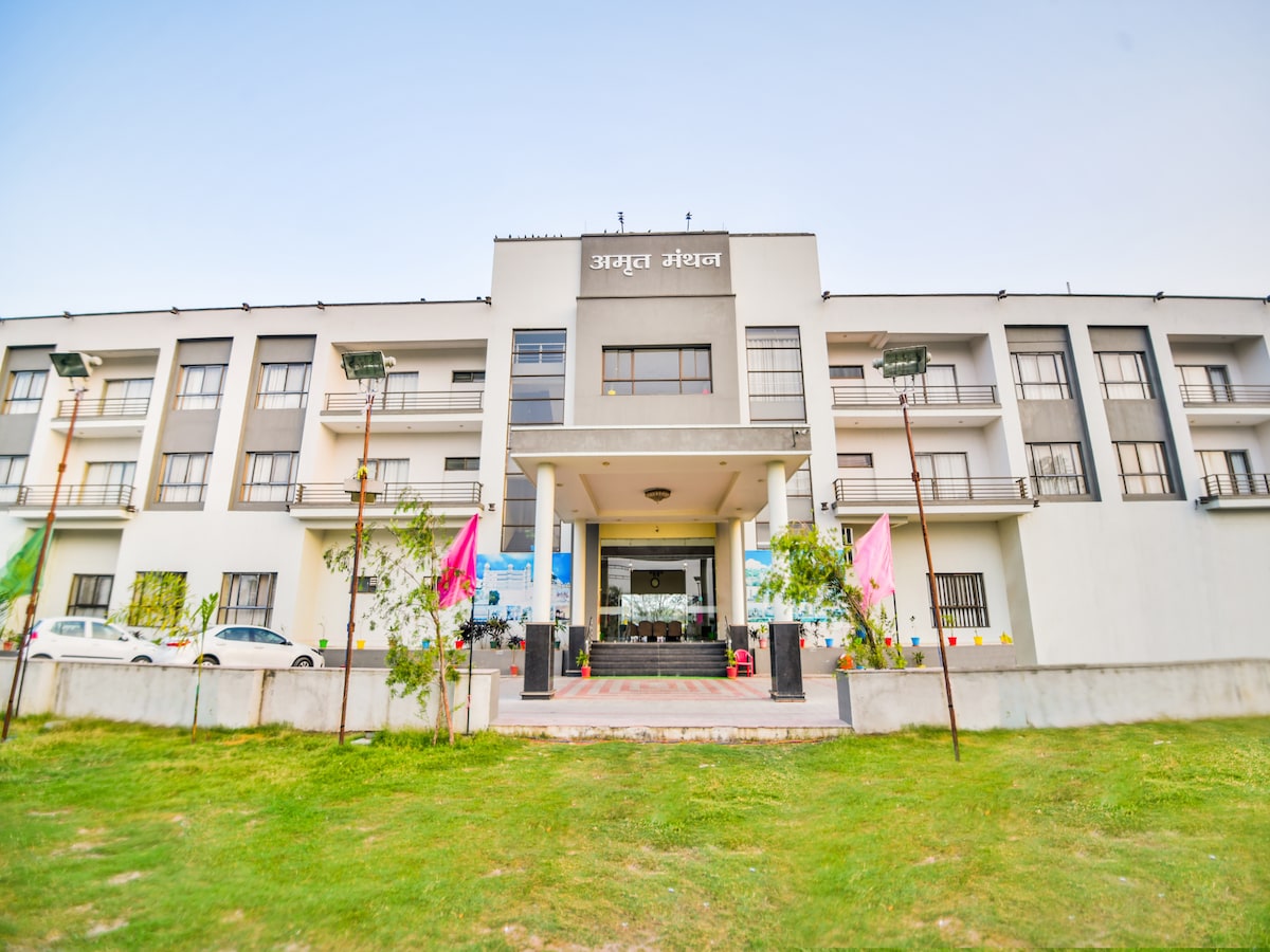 The hotel exterior features a modern design with a spacious entrance. Colorful flags are displayed alongside neatly maintained greenery, while large windows provide ample natural light. The parking area accommodates guest vehicles, adding convenience for visitors.