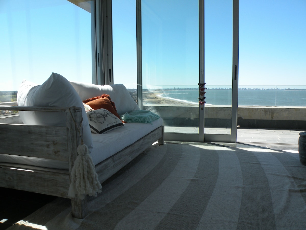 A comfortable seating area is presented with a white daybed adorned with colorful cushions. Large glass doors offer an unobstructed view of the ocean, while a neutral-toned area rug complements the light and airy space.