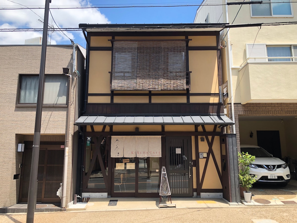 The exterior of the guesthouse is showcased, featuring a traditional Japanese architectural style with wooden beams and a slatted window covering. A welcoming entrance is emphasized by a sign displaying the name and a pathway leading to the door. Surrounding structures are visible in a blend of modern and traditional styles.