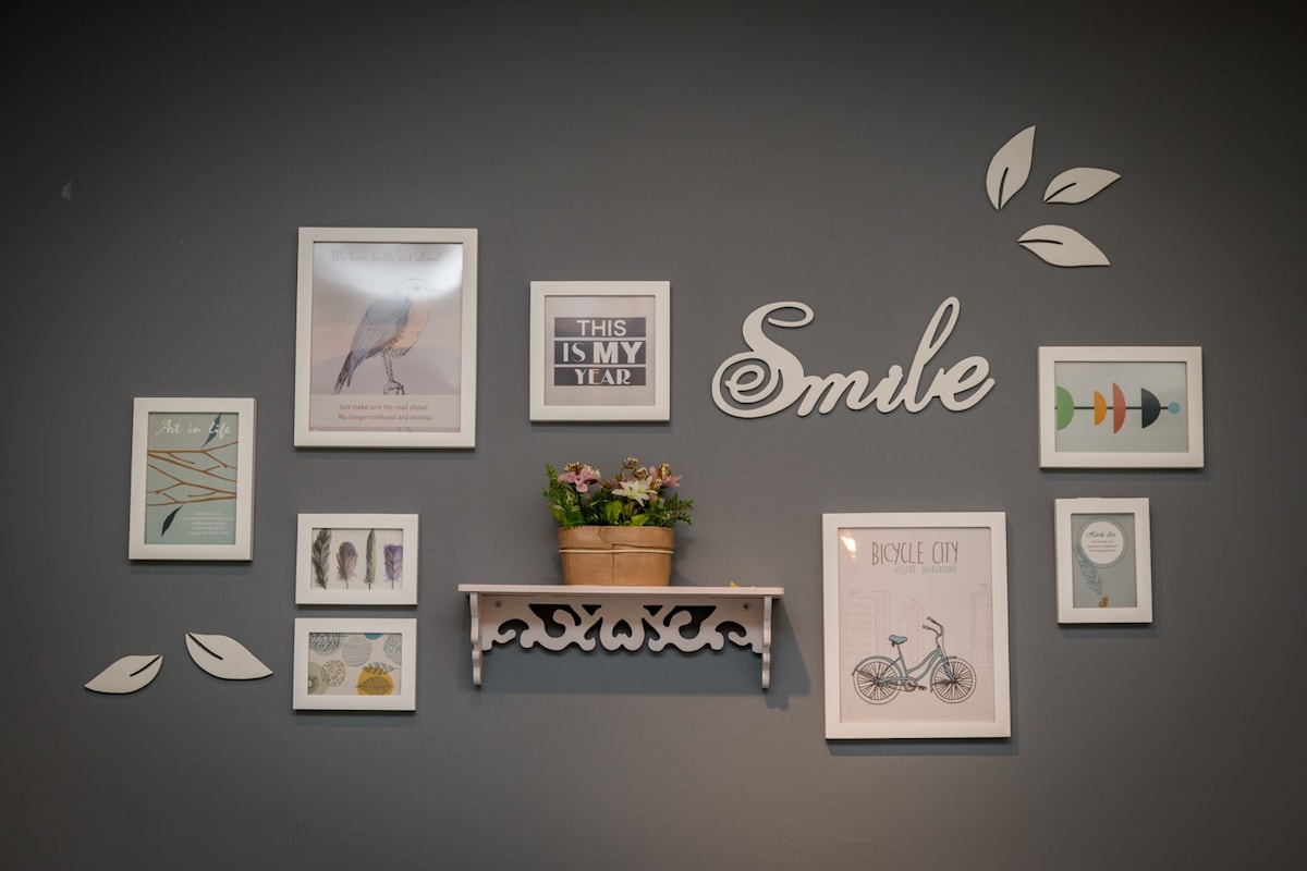 A gallery wall is displayed, featuring framed art prints and inspirational messages against a dark wall. A small shelf holds a potted plant, enhancing the decor with a touch of greenery. The arrangement includes diverse artwork and playful elements.