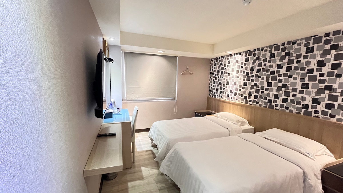A modern and tidy room features two spacious beds, a wall-mounted television, and a compact desk area. Natural light enters through a window adorned with a roller shade, while decorative wall accents add texture to the space.