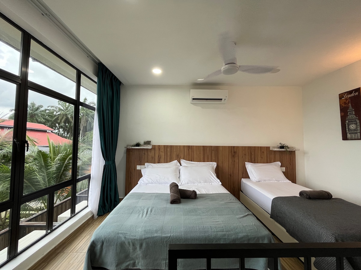 A bright bedroom features two spacious beds with fresh linens. Natural light filters through large windows adorned with green curtains, providing a view of the surrounding greenery. A ceiling fan is visible, adding to the comfortable atmosphere.