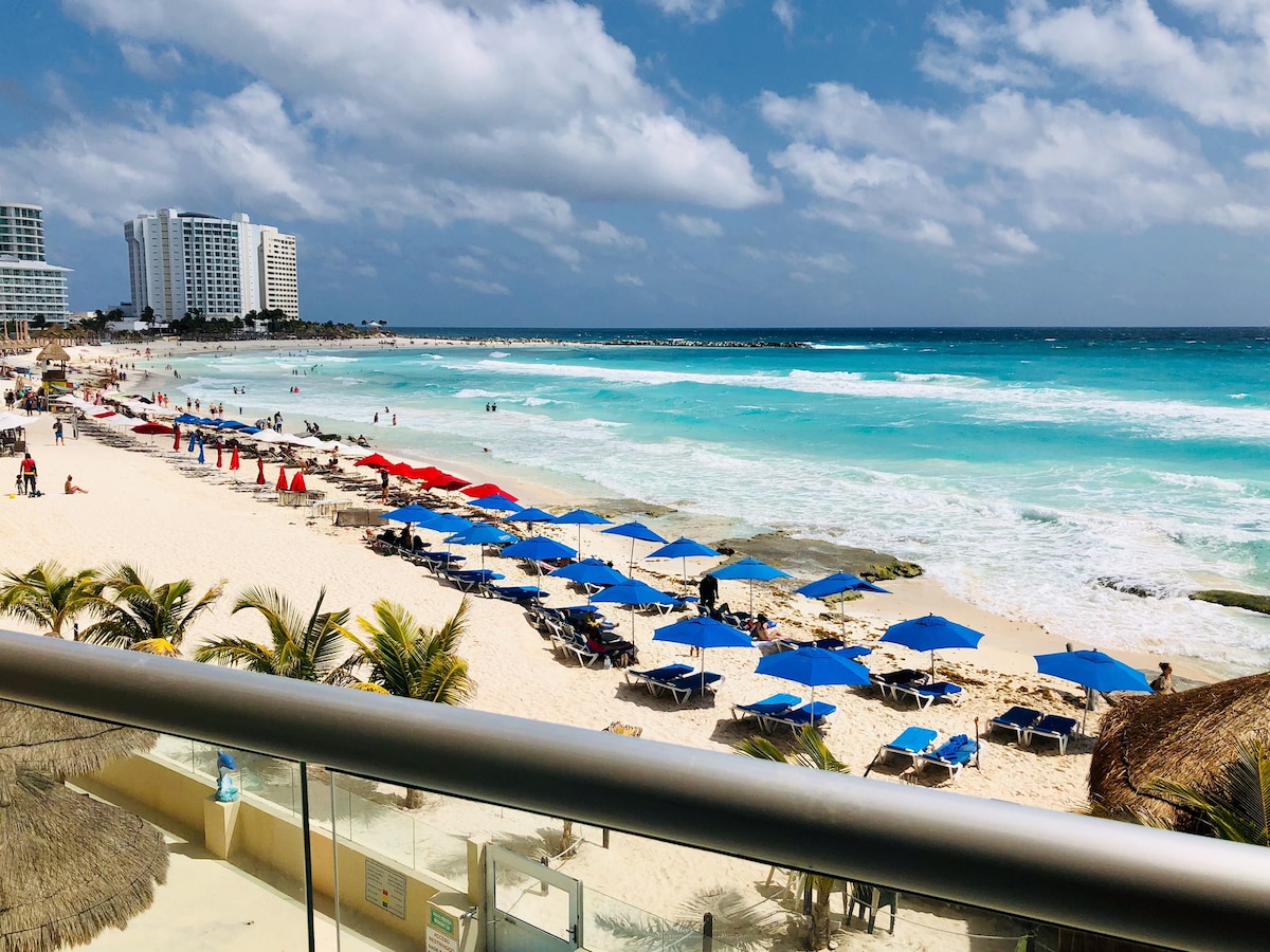 A view of the vibrant beach scene from the studio balcony, featuring clusters of blue umbrellas and sun loungers on the sandy shore. The turquoise waves gently lap against the coastline under a partly cloudy sky, while buildings in the background define the horizon.