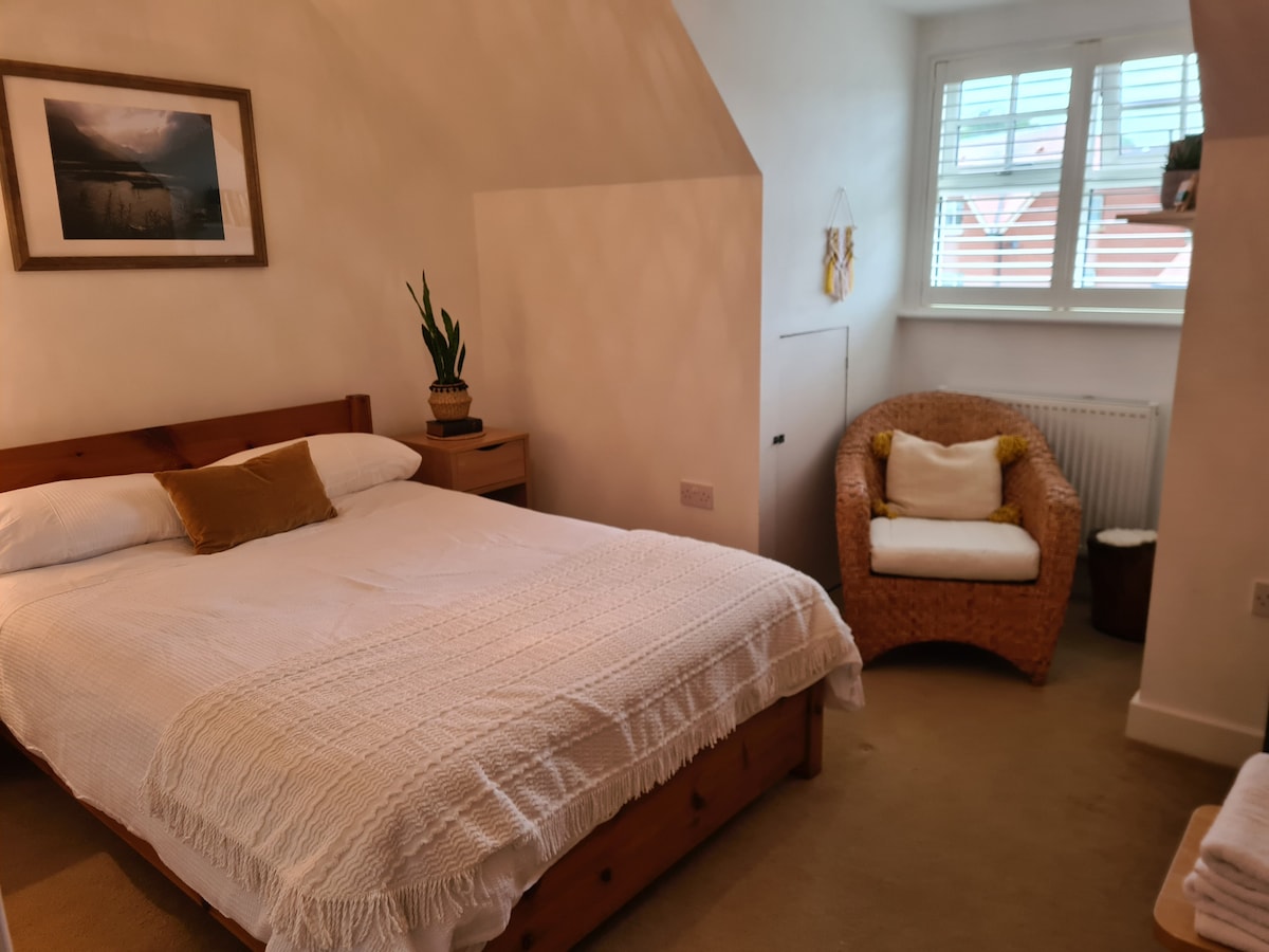 A private room features a spacious bed adorned with a white knitted throw and a decorative pillow. A comfortable rattan chair adds warmth to the space, while a small wooden bedside table holds a plant and framed artwork decorates the wall.