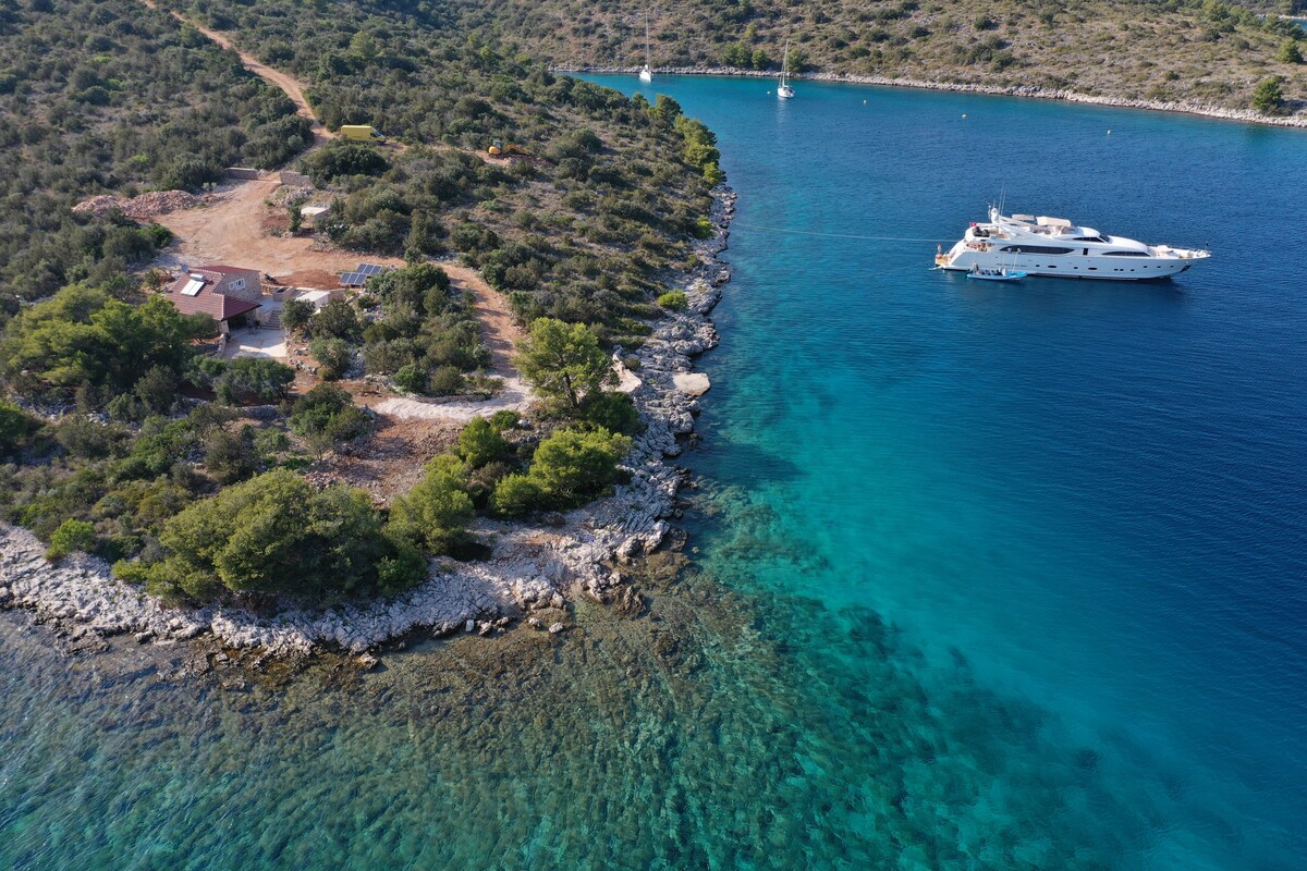 An aerial view captures the seafront property alongside clear turquoise waters. A private boat is moored nearby, while lush greenery and rocky shoreline frame the image, highlighting the secluded nature of the bay. The coastline reveals a blend of land and water, inviting exploration.