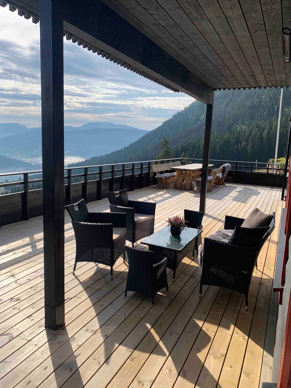 A spacious terrace with wooden flooring features a seating arrangement of four black wicker chairs and a glass coffee table, surrounded by stunning mountain views. A large dining table is positioned toward the back, inviting outdoor meals amid a serene landscape.