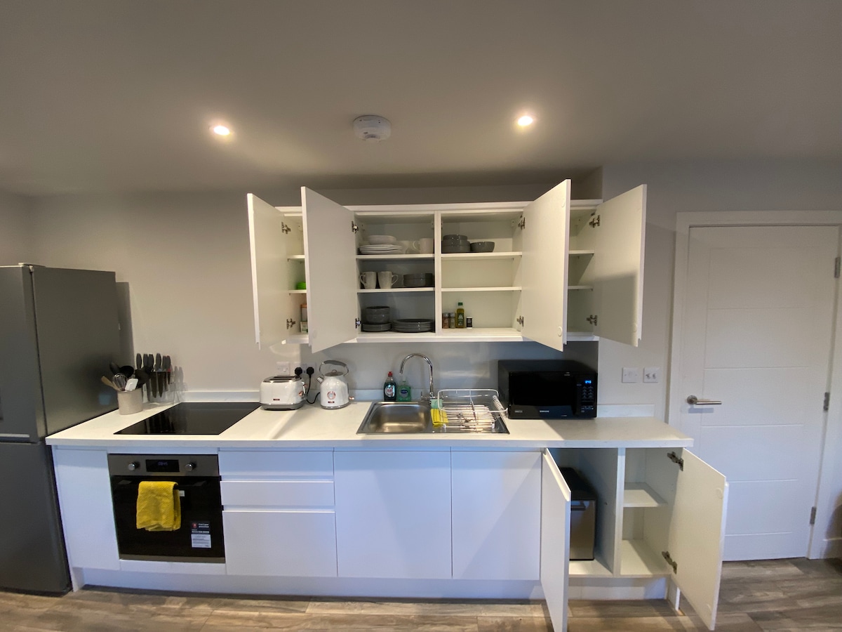 A modern kitchen features white cabinetry with open doors showcasing organized dishware. The countertop includes a double sink, stove, and microwave. A refrigerator is located to the left, while a small appliance area is visible. Storage space below is also accessible.