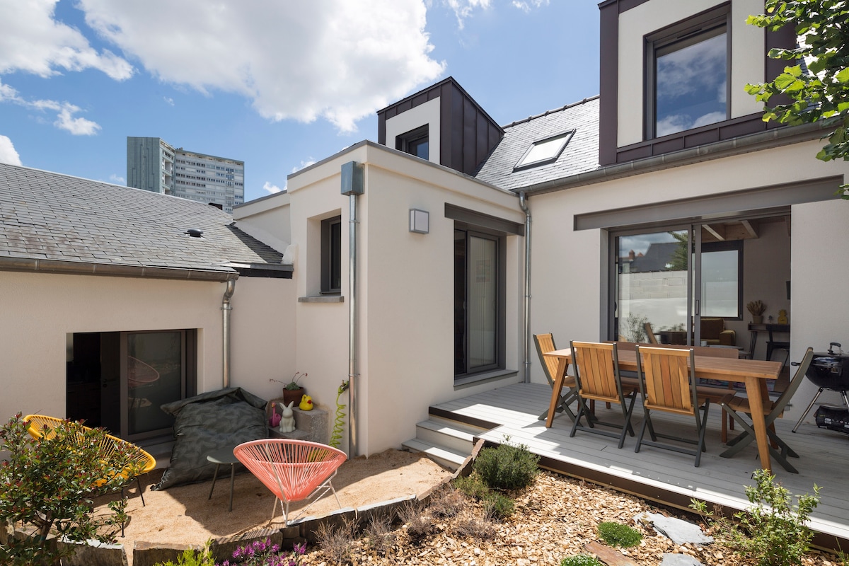 The exterior of the house is showcased, featuring a modern design with large windows and a spacious terrace. A wooden dining set is visible on the deck, surrounded by a small garden area with gravel and plants. The urban backdrop includes nearby buildings, contrasting with the outdoor tranquility.