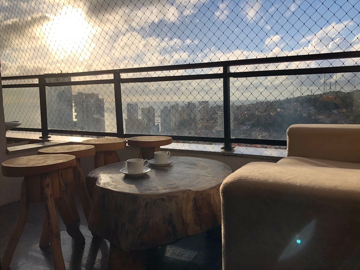A cozy balcony setting features a wooden table and three stools, with two coffee cups placed atop the table. A city view is visible through a mesh screening, illuminated by soft morning sunlight.