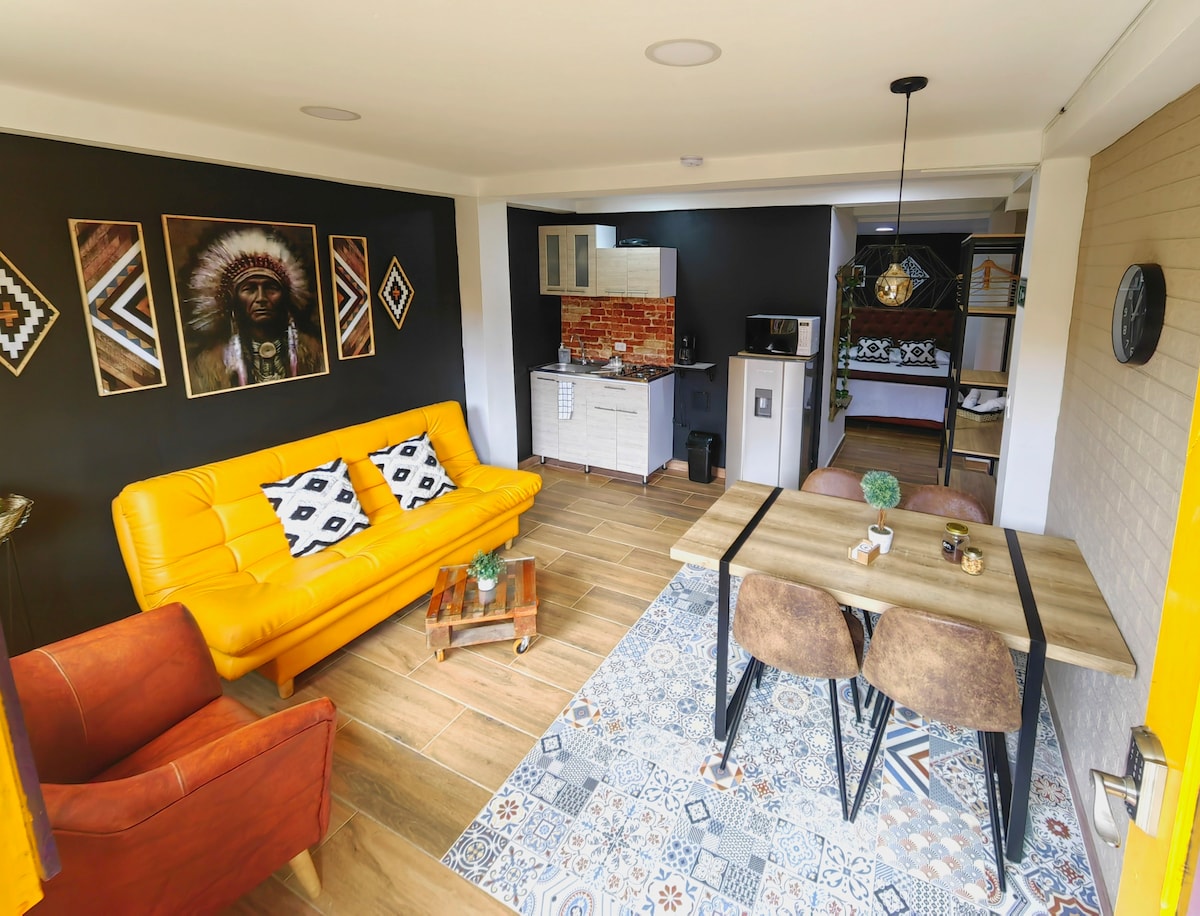 The open living area features a vibrant yellow sofa and an orange armchair, paired with a dining table for four. A modern kitchenette is visible, showcasing essential appliances. Unique wall art adds character to the space, enhancing the overall design.