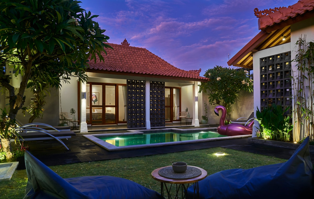 The serene outdoor area features a private pool accompanied by vibrant green landscaping. Two comfortable seating areas are positioned on the lawn, offering relaxation space, while traditional architecture with a striking red roof frames the inviting atmosphere.