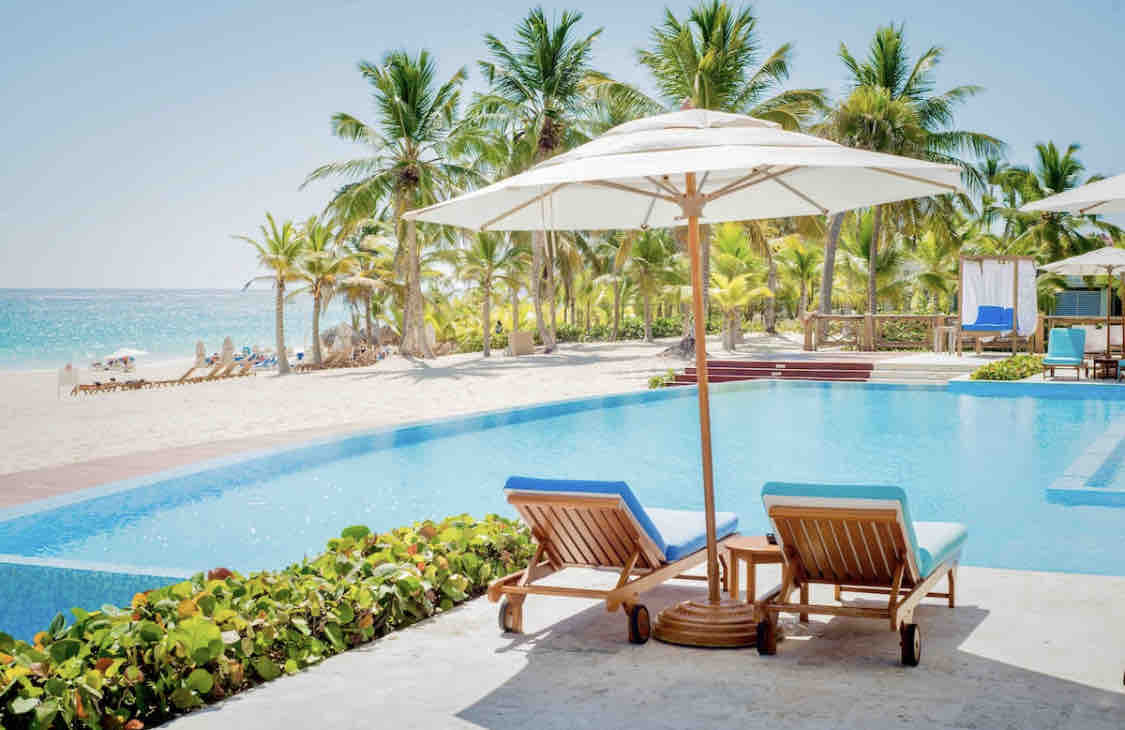 A serene infinity pool is framed by lush palm trees and vibrant greenery. Two blue lounge chairs sit under a white umbrella, inviting relaxation by the water's edge with a view extending towards the beach and ocean in the background.