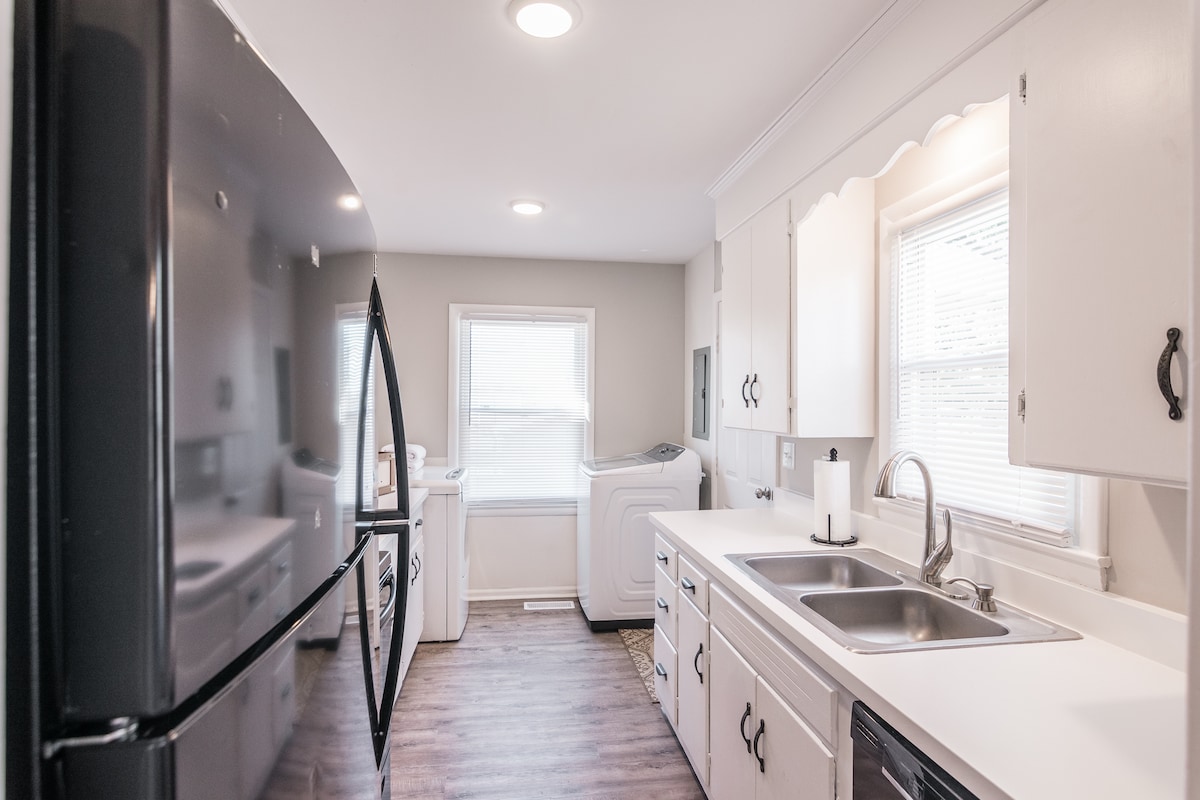 A well-organized kitchen features white cabinetry and stainless steel appliances. A dual sink is situated between the windows, allowing natural light to brighten the space. The black refrigerator stands opposite a washer and dryer, providing convenience for guests.