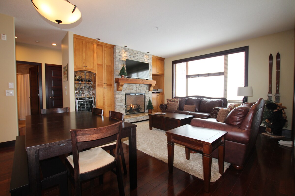 A welcoming living area features a cozy fireplace surrounded by stone, with a large window allowing natural light to fill the space. Comfortable seating is arranged invitingly, complemented by a dining table nearby. Ski gear is subtly displayed, adding to the vacation atmosphere.