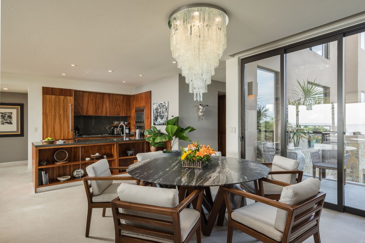 A dining area features a round stone table surrounded by four wooden chairs with soft cushions. A stunning crystal chandelier hangs overhead, providing warm light. In the background, a modern kitchen with wooden cabinetry and a coffee maker is visible, along with lush greenery outside through expansive glass doors.