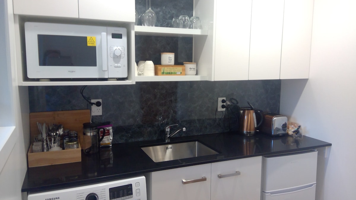 The kitchenette features modern appliances, including a microwave, kettle, and toaster. Black countertops provide a sleek surface, while a well-organized shelf holds glassware. A washing machine is integrated into the cabinetry, ensuring convenience for guests.