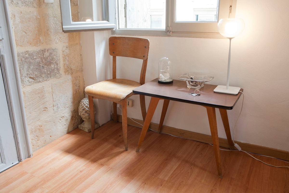 A simple corner is highlighted by a wooden chair beside a small table. The table features a decorative glass bowl and a sleek lamp, providing soft illumination. Natural light enters through the nearby window, enhancing the light wood flooring and the stone wall's texture.