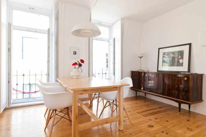 A bright dining area features a wooden table surrounded by white chairs, enhanced by natural light from two open doors. A decorative vase with flowers sits in the center of the table, while a wooden cabinet with sleek design is positioned against the wall.