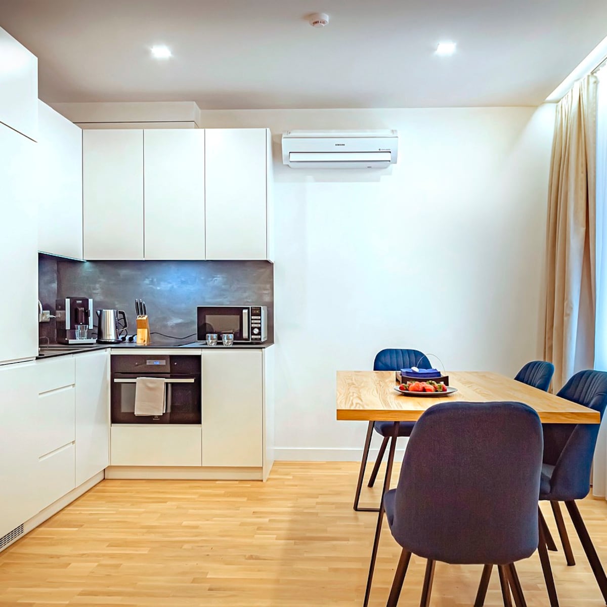 A modern kitchen features sleek white cabinetry and a dark countertop, equipped with essential appliances including a coffee maker and microwave. A wooden dining table is surrounded by blue chairs, and natural light streams in from a window draped with light curtains.