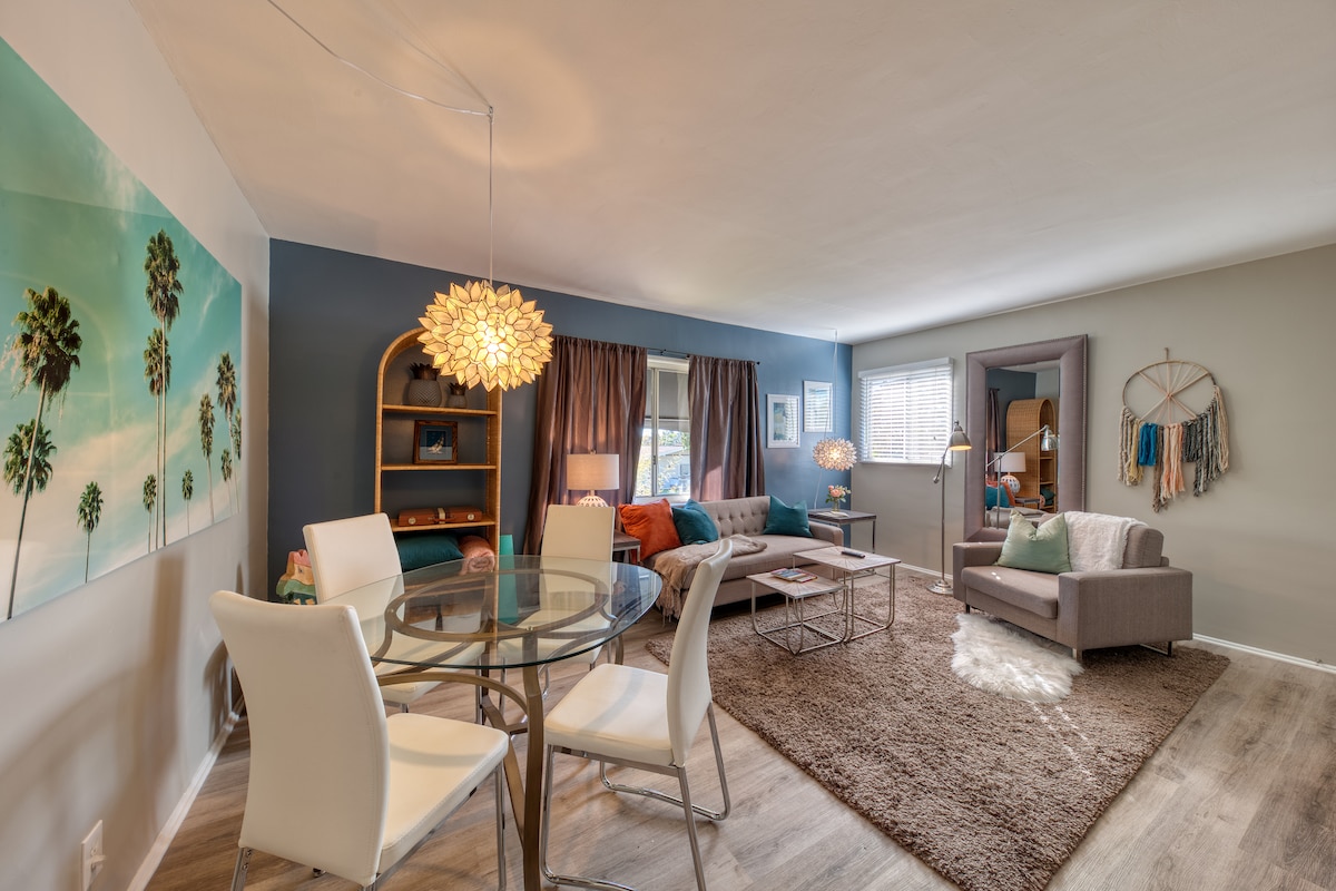 A bright living area is presented with a circular glass dining table surrounded by four white chairs. A plush area rug covers the floor, and colorful cushions accent a light grey sofa. Large windows are dressed with dark curtains, allowing natural light to fill the space.