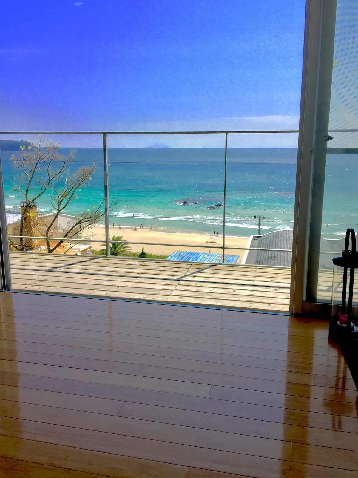A spacious indoor area presents a large glass door that opens to a stunning view of the beach and ocean. The clear blue water and sandy shore are visible, framed by a wooden deck. Natural light fills the room, enhancing its serene atmosphere.