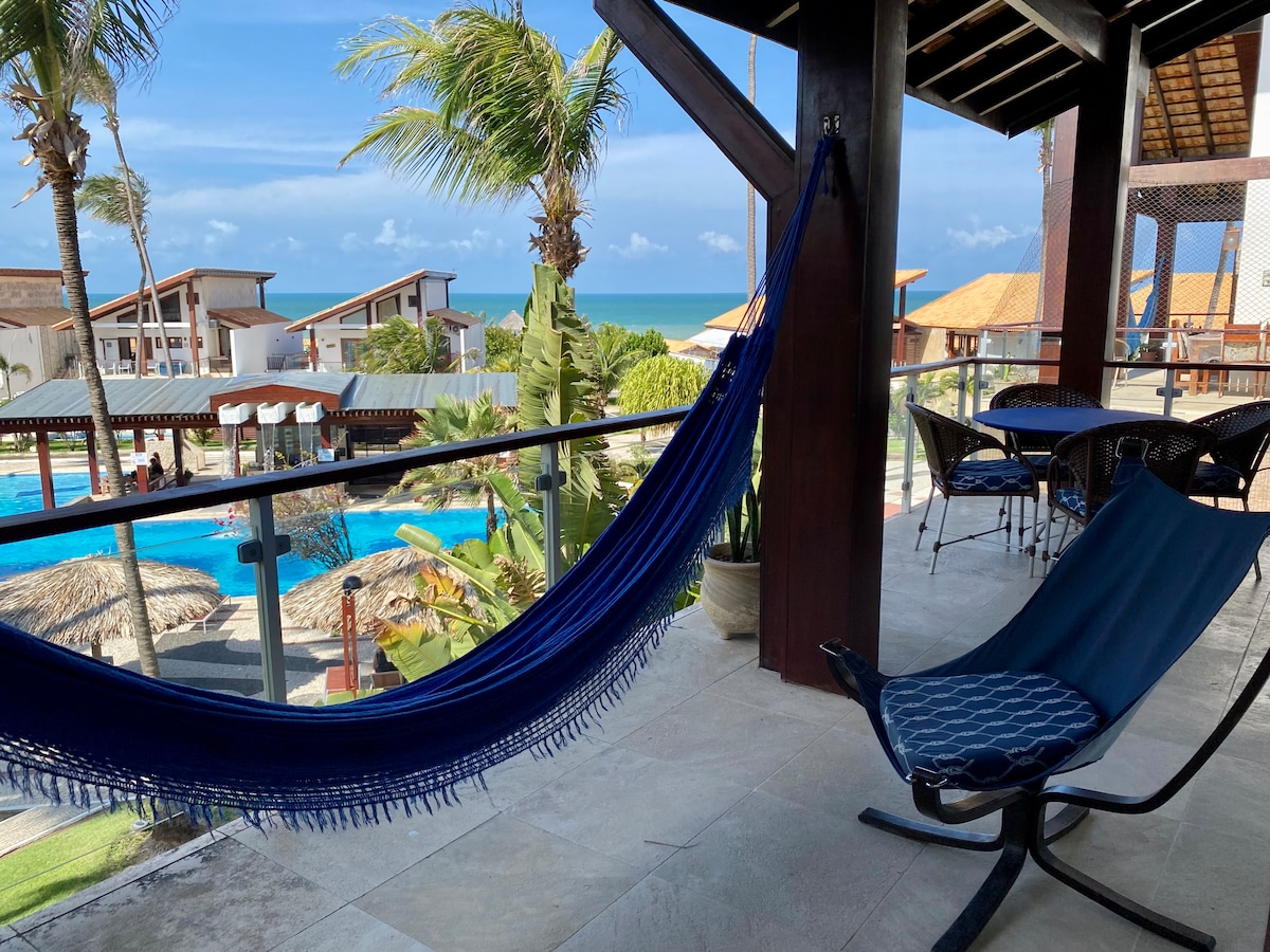 A hammock hangs gracefully on a balcony, overlooking a pool area surrounded by palm trees. The sea is visible in the distance, and comfortable chairs are positioned nearby, offering a relaxed space to enjoy the view.
