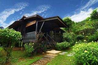 A traditional Malay house on stilts is surrounded by lush greenery and flowering plants. Steps lead to a welcoming veranda, offering a view of the vibrant garden. The house's dark wood exterior contrasts with the bright blue sky, creating a serene atmosphere.