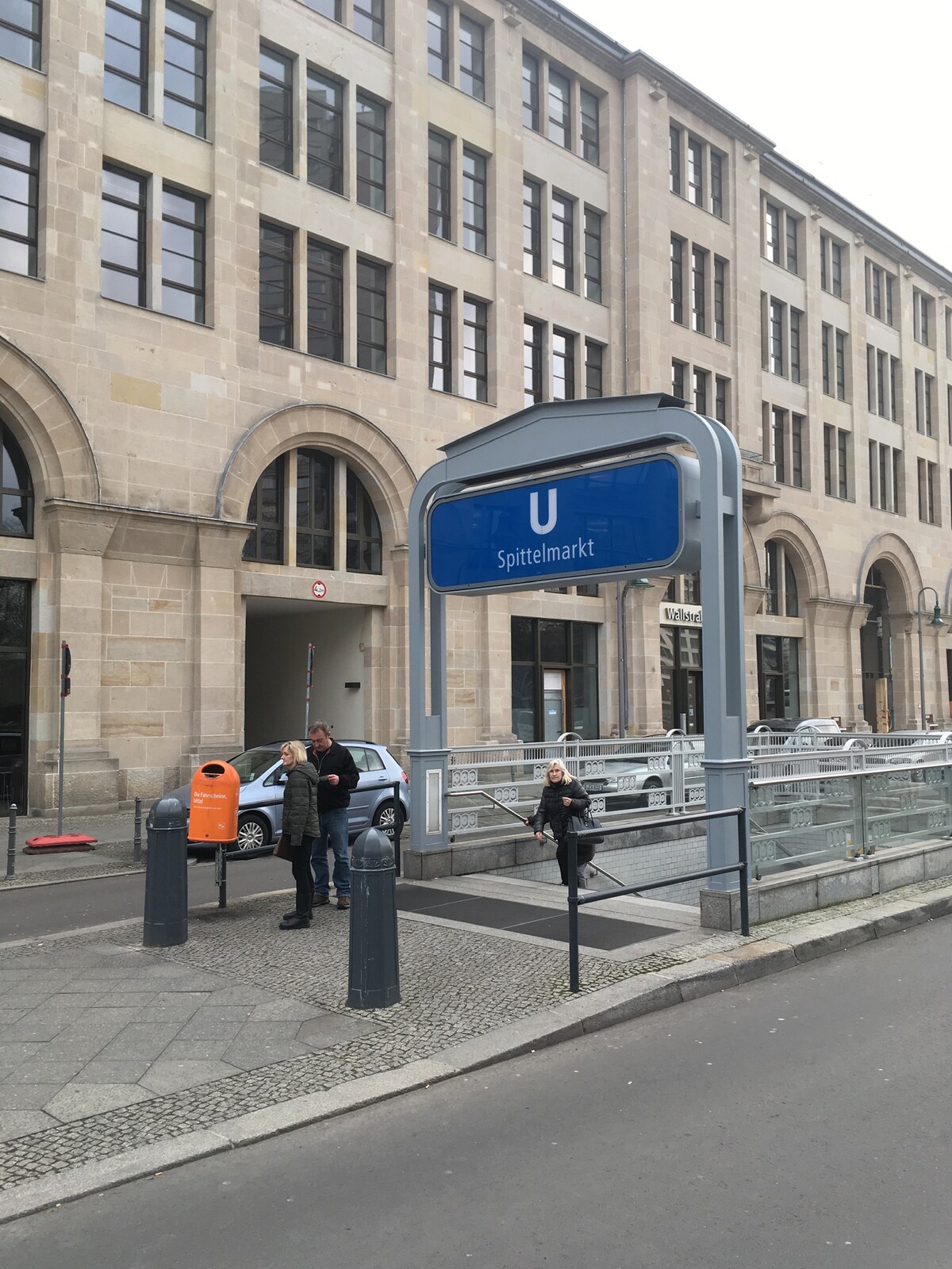 The image showcases the entrance to the Spittelmarkt underground station. A large blue sign indicating 'U' is prominently displayed. The surrounding area features a modern stone building with multiple windows, and people are seen near the entrance, engaging in various activities.