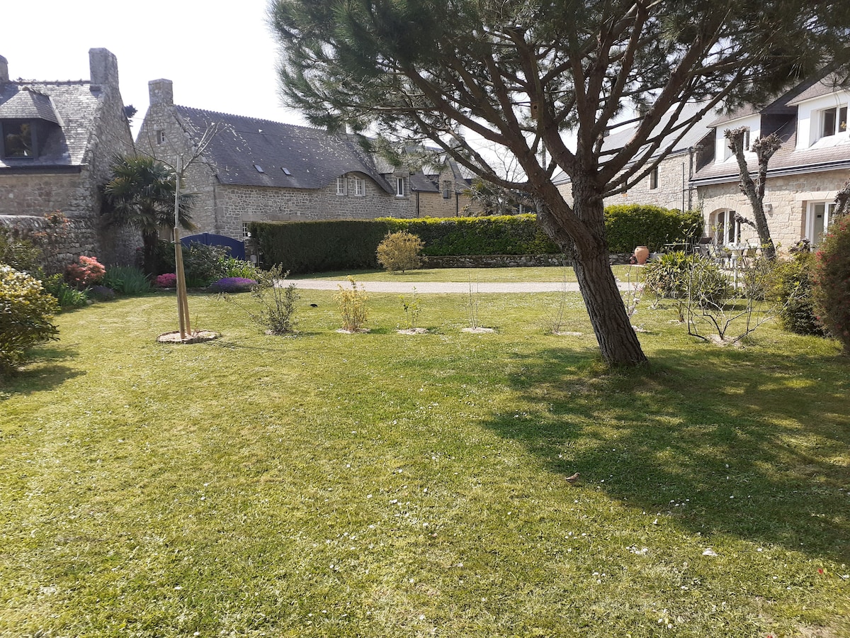 A well-maintained garden area is shown with a neatly mowed lawn. A large tree provides shade, and various shrubs and plants are distributed throughout the space. The backdrop features several traditional stone houses, highlighting the peaceful surroundings of the property.