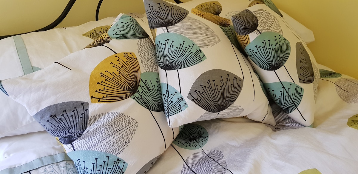 A neatly made double bed is adorned with a light-colored duvet featuring a botanical print. Several decorative pillows in varying colors and patterns are arranged at the head of the bed, adding visual interest to the room's serene yellow walls.